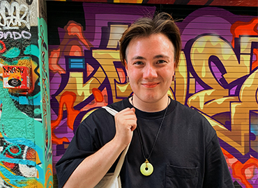 picture of Thomas holding a canvas bag, with a wall of graffiti in the background