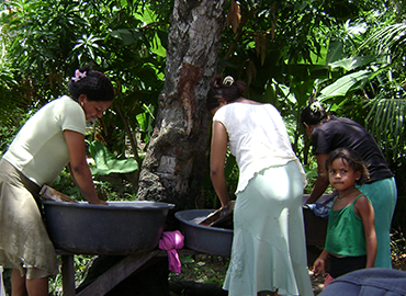 Two women bending over wheelbarrows in a forested area. A child stands in front of them, looking at the camera.